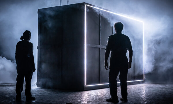 Two people stand still facing a very large crate. There are strips of bright white light coming from the border of the crate