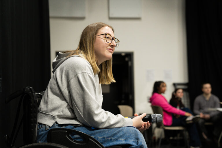 The Access Manifesto: A Pioneering Movement Set to Transform the Arts A young woman (Maisy Gordon) with blonde hair and glasses sits in a wheelchair, holding a microphone. She is wearing a grey hoodie and blue jeans, looking slightly to her right. In the background, other people are visible, out of focus.