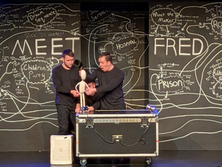 Two men manipulate a puppet on stage in front of a blackboard with "MEET FRED" and various words and diagrams written in white chalk.