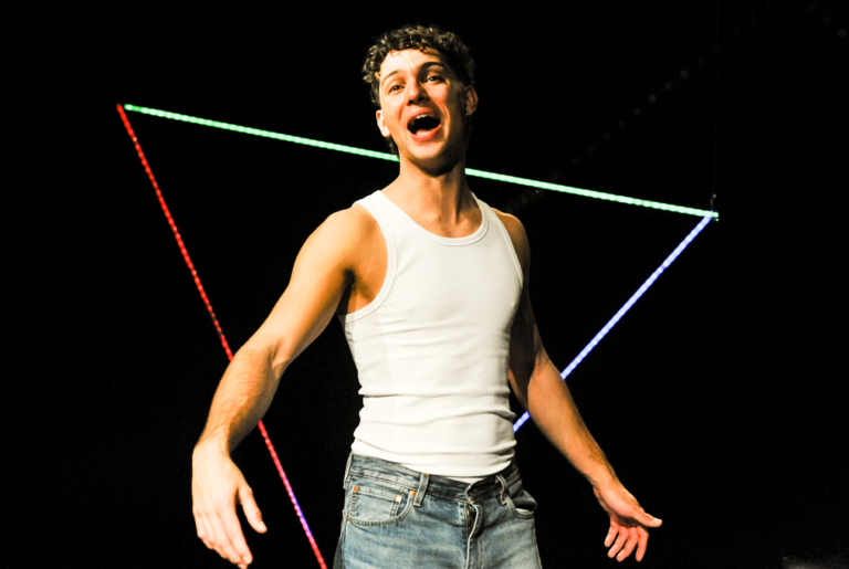 Alt text: A young man wearing a white tank top and blue jeans stands against a dark background, smiling with his mouth open as if singing or speaking. Colorful neon light strips form triangular shapes behind him, creating a vibrant, stage-like atmosphere.