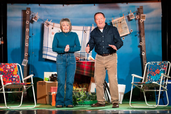 Someone’s Knockin’ At The Door – Lemon Tree, Aberdeen a middle aged man and woman stand in front of a blue background with deckchairs behind them