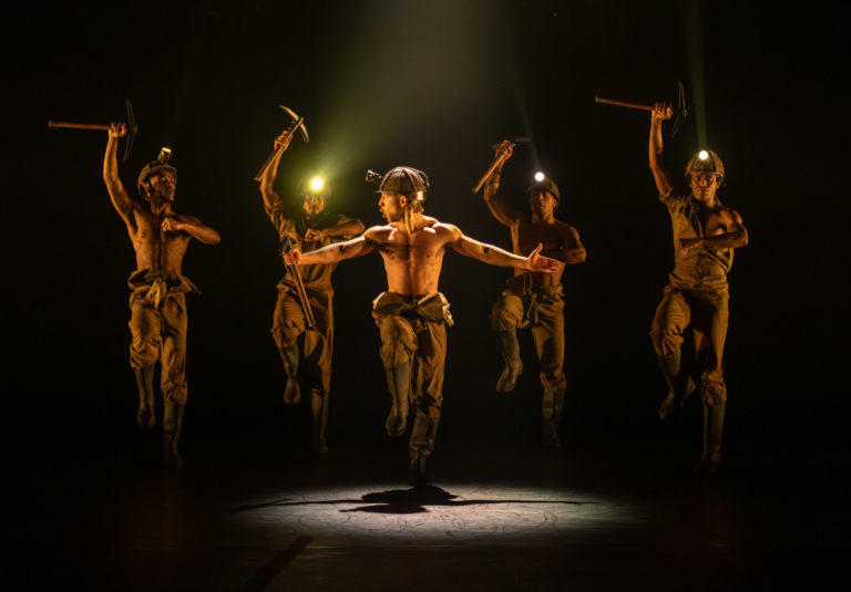 Five dancers on a dark stage perform a synchronized routine while dressed as miners, wearing helmets with headlamps and holding pickaxes. The central dancer, shirtless, stands slightly forward with arms extended, while the others leap or pose mid-motion around him under dramatic, warm spotlighting.