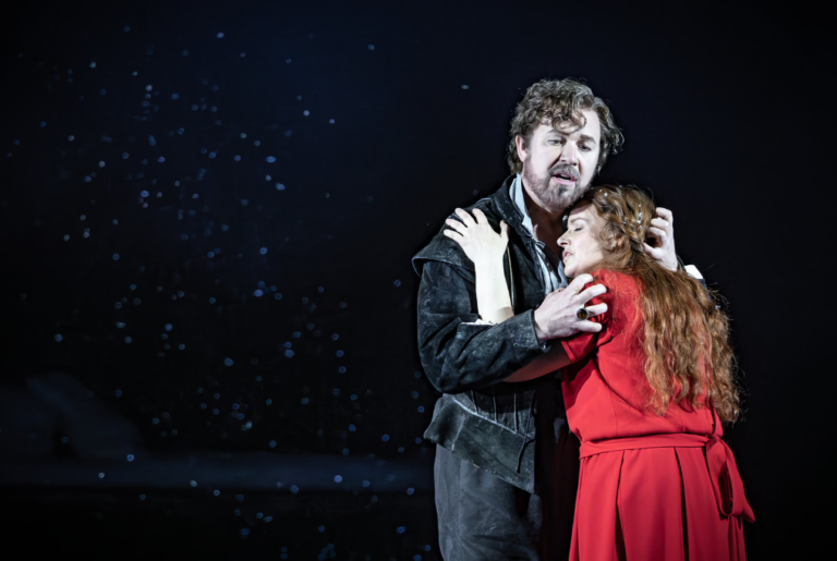 A dramatic stage scene shows a man in dark, weathered clothing holding and comforting a woman in a bright red dress. They embrace closely, the woman resting her head against his chest while he looks outward with a pained expression. The background is dark with soft, scattered light specks, emphasizing the emotional intensity of the moment.