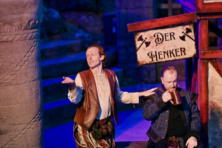 Two actors in period costumes perform on stage; one gestures expressively while the other drinks from a mug beside a sign reading "Der Henker.