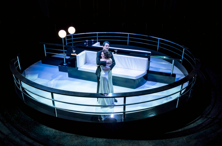 Private Lives- Royal Exchange, Manchester A couple embraces at the center of a stylized ship deck set, surrounded by curved railings and soft glowing lights, evoking a dramatic, theatrical scene reminiscent of a luxury ocean liner at night.