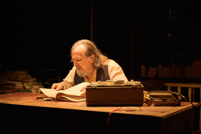 Gary Oldman looking at an open book, sat at a desk.