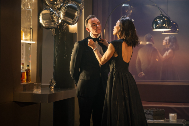 A woman in a black dress adjusts a man’s bow tie as he stands in a tuxedo at an elegant indoor celebration, with silver balloons, a bar shelf with bottles, and a softly lit mirror reflecting the scene in the background.