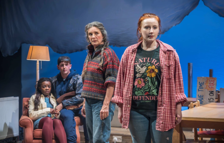 Atlantis – Theatr Clwyd, Mold Four actors stand and sit on a stage set resembling a living room. In the foreground, a young red-haired woman in a pink plaid shirt and graphic T-shirt stands beside an older woman in a patterned sweater, both looking ahead with serious expressions. Behind them, a young Black woman sits in an armchair while a young man sits nearby. A floor lamp, table, and chairs complete the domestic setting, while a large blue backdrop overhead creates a dramatic, theatrical atmosphere.