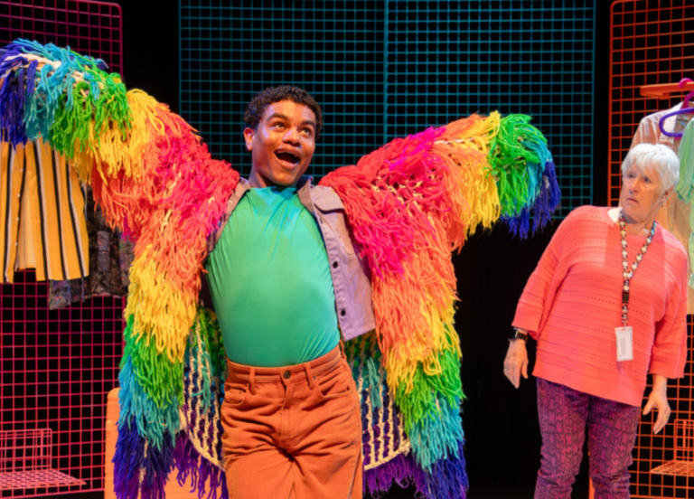 A person on a brightly lit stage poses with arms outstretched while wearing a dramatic rainbow-colored fringed coat that spreads like wings. They have a turquoise shirt and orange trousers, and their joyful expression suggests a theatrical performance. An older woman in a pink top and patterned purple trousers stands nearby looking toward them. The set features colorful grid panels and clothing racks displaying garments in the background.