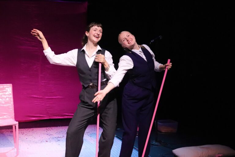 Two performers stand on a dimly lit stage with a pink backdrop, smiling and posing with long pink poles. Both wear white button-down shirts, dark vests, and tailored trousers. One performer gestures outward while the other leans in, creating a playful, theatrical pose. A white chair sits to the left, and a pillow is visible on the floor at the right edge of the stage. Pink and purple lighting casts a vibrant glow over the scene.