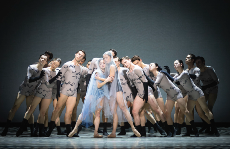 A contemporary ballet ensemble performs on a dimly lit stage. At the center, two dancers embrace, one draped in a translucent pale-blue veil and tulle costume, while a group of dancers in matching gray bodysuits with black gloves and socks surround them in a close formation. The ensemble leans inward with heads tilted and eyes closed or downcast, creating a sense of intimacy, support, and emotional tension under a single overhead spotlight.