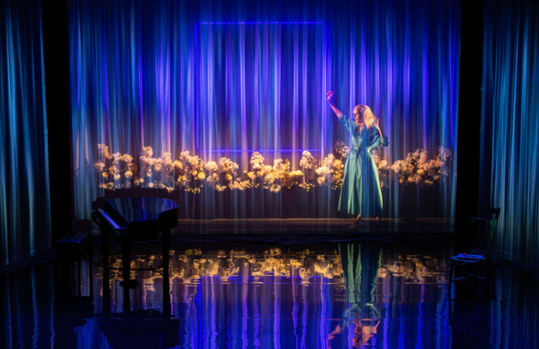 Mrs Dalloway – Storyhouse, Chester A theatrical stage glows in deep blue and violet light. A woman in a flowing light-blue dress stands near the center-right, raising one arm as if dancing or performing. Behind her, sheer curtains are illuminated by vertical bands of light, with a row of pale flowers stretching across the stage. A black grand piano sits in shadow at the front left, while the glossy floor reflects the lights, flowers, and performer, creating a dreamlike atmosphere.