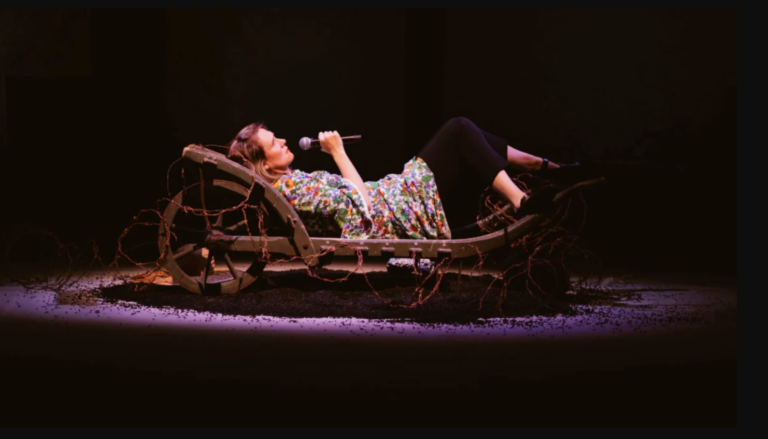 Bryony Kimmings: Bog Witch – Storyhouse, Chester Bryony Kimmings reclines on a weathered wooden cart at centre stage, holding a handheld microphone close to her mouth as if singing or delivering a monologue. She wears a vibrant floral dress over black leggings and black shoes, illuminated by a warm spotlight against a dark, minimalist backdrop. Twisted branches and exposed roots entwine the cart and spread across the stage floor, which is scattered with dark soil, creating an earthy, atmospheric setting that evokes themes of nature, folklore and isolation.