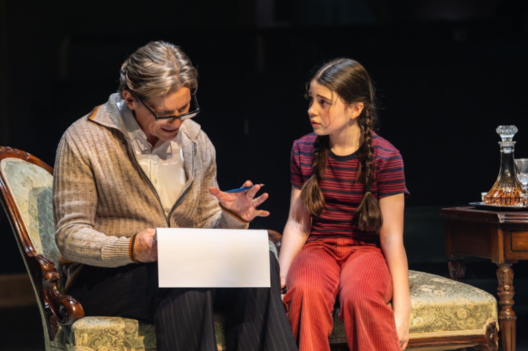 An older adult wearing glasses and a beige cardigan sits beside a young girl with braided hair on a vintage sofa under stage lighting. The older adult gestures while looking at a sheet of paper or drawing pad on their lap, and the girl watches with a thoughtful, slightly concerned expression. A wooden side table with a crystal decanter and glasses stands nearby against a dark theatrical backdrop.
