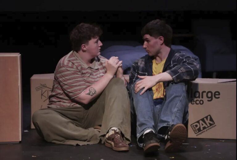Lovemaking To An Inappropriate Soundtrack – 53two, Manchester Two performers sit on the floor of a dark theatre stage, facing each other in an intimate conversation. They lean against stacked moving boxes with a bed visible in the background, suggesting a recent move. One performer clasps the other's hands while both maintain serious, attentive expressions, creating a quiet, emotional moment within the stage set.
