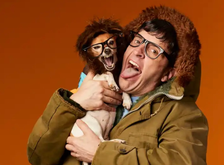 A person wearing oversized glasses and a fur-lined hooded jacket holds a small dog dressed in a shaggy wig and matching glasses. Both are making exaggerated, playful expressions with their mouths open against a bright orange studio background, creating a humorous and quirky portrait.