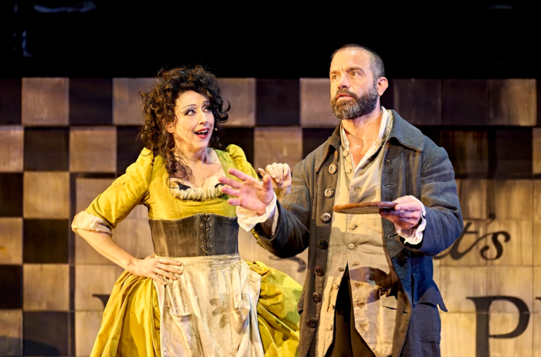 Sweeney Todd : The Demon Barber of Fleet Street – Birmingham REP Ramin Karimloo as Sweeney Todd and Meow Meow as Mrs Lovett perform on stage in Sweeney Todd. Mrs Lovett, dressed in a bright yellow period gown and apron, looks at Sweeney Todd with an animated expression as he gestures dramatically while holding a pie plate. The scene is set against a large checkerboard-style backdrop under warm theatrical lighting.