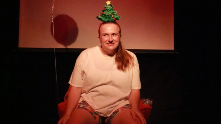 A performer sits on stage wearing a white T-shirt, colourful patterned shorts and a Christmas tree headband, smiling towards the audience. A warm red-lit backdrop and stage screen are visible behind them.