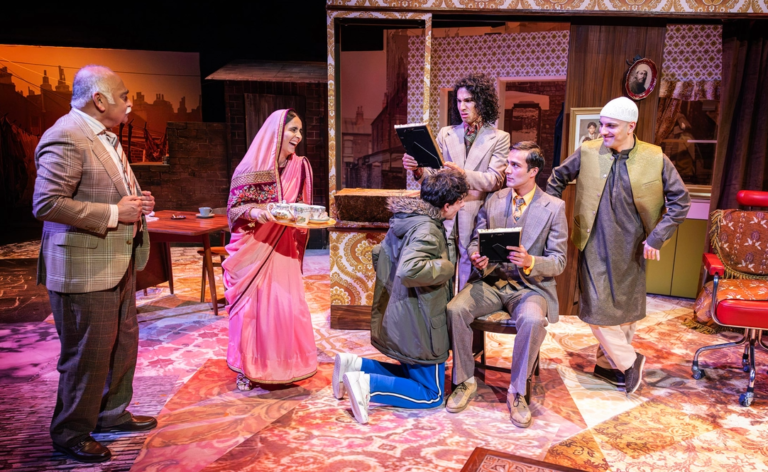 East Is East- Octagon Theatre, Bolton A cast of six performers gather on stage in a richly decorated living room set. A woman in a pink sari carries a tea tray, while others in period-style clothing interact around a seated man and a child kneeling in front of him.