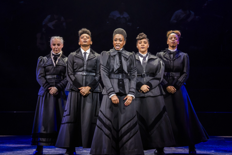 Sylvia : A Musical Revolution – The Lowry, Salford Five performers stand in a powerful formation on stage in Sylvia, wearing coordinated black Victorian-inspired dresses with fitted jackets and long skirts. The central performer appears to be singing or delivering a passionate speech, flanked by four women standing defiantly. Dramatic stage lighting highlights the cast against a dark background, with musicians faintly visible behind them. Photograph by Tristram Kenton.