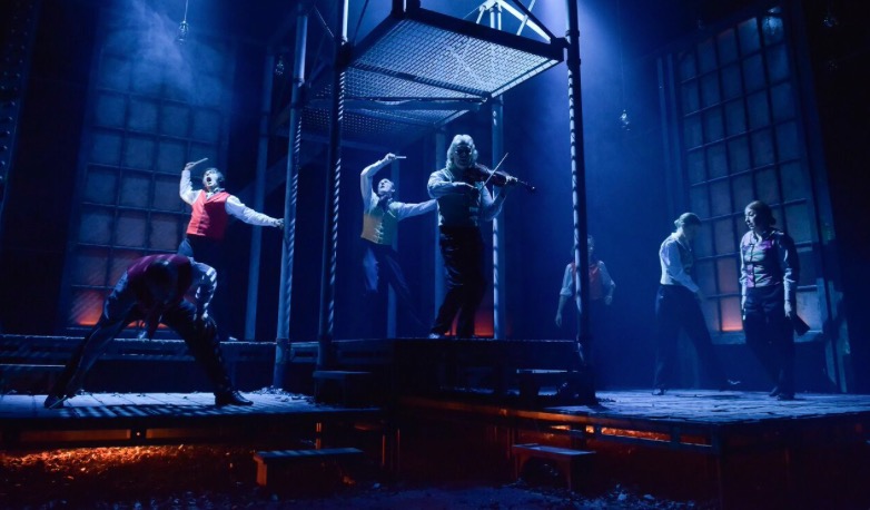 The cast are silhouetted against a set of iron girders bathed in blue light