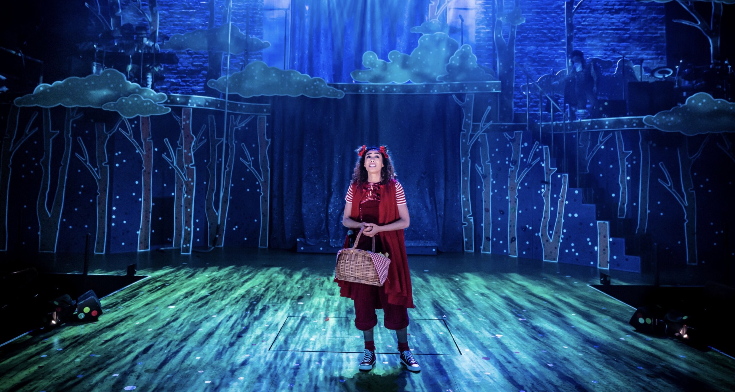In a dark shadowed woodland, Red Riding Hood (Paisley Reid) wears red hood and carrying a basket.
