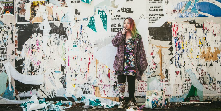 woman smoking stands in front of a wall of torn posters